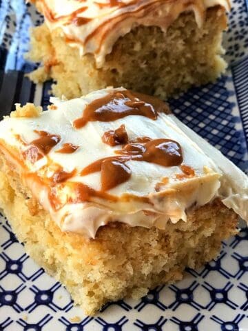 Two square slices of banana caramel cake topped with white frosting and a caramel swirl on a patterned blue and white plate.