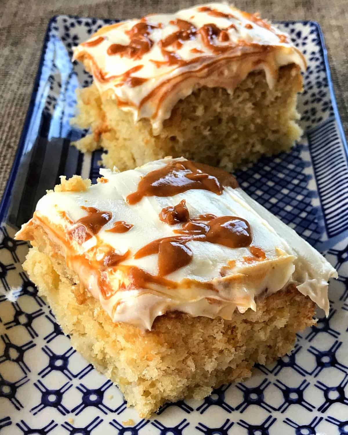 Two squares of banana caramel cake topped with white frosting and a swirl on a patterned blue and white plate.