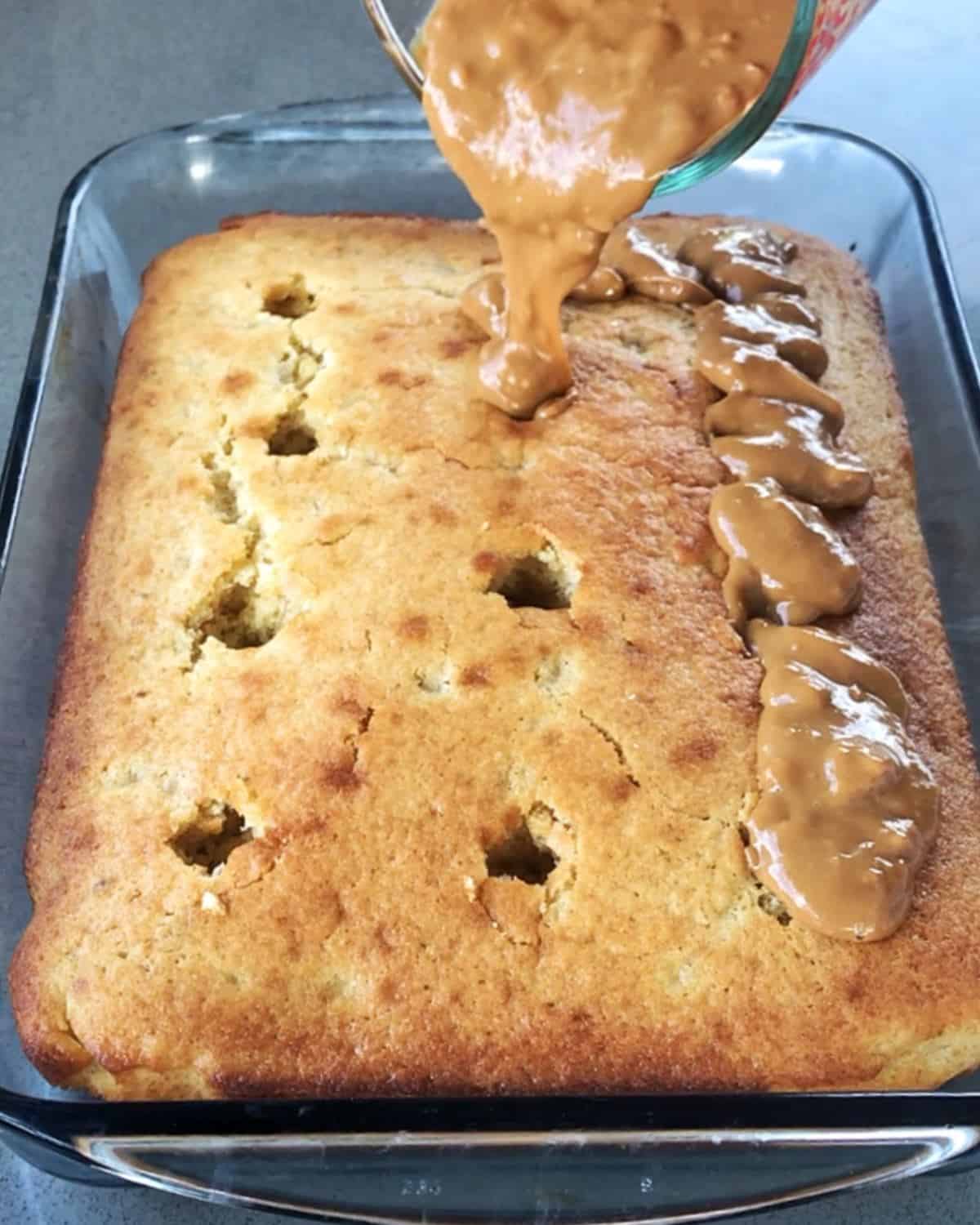 Creamy peanut butter being poured over a banana caramel sheet cake with holes poked in the surface.