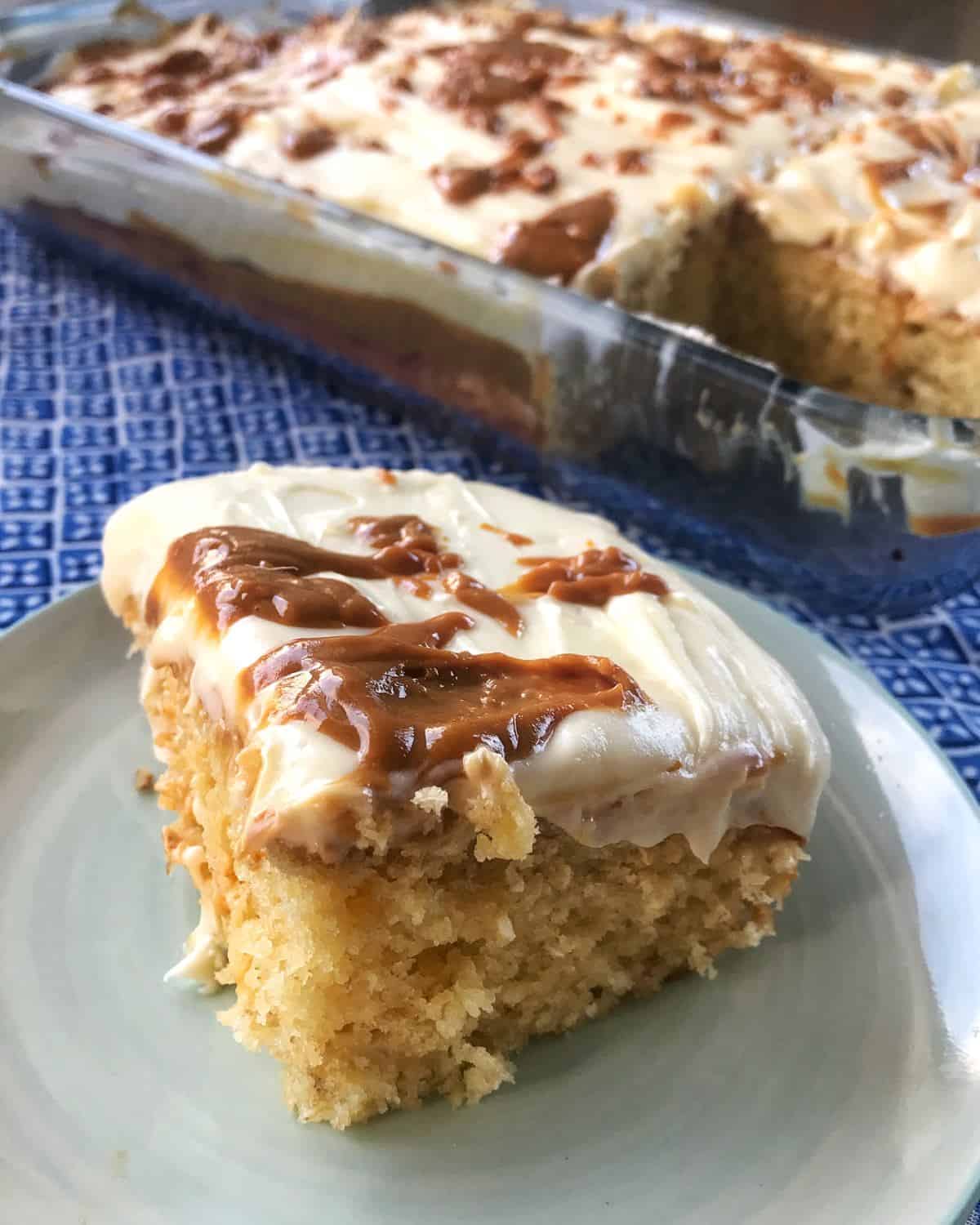 A slice of banana caramel cake topped with white frosting and swirls of caramel on a small plate, with a glass pan of the same cake in the background.