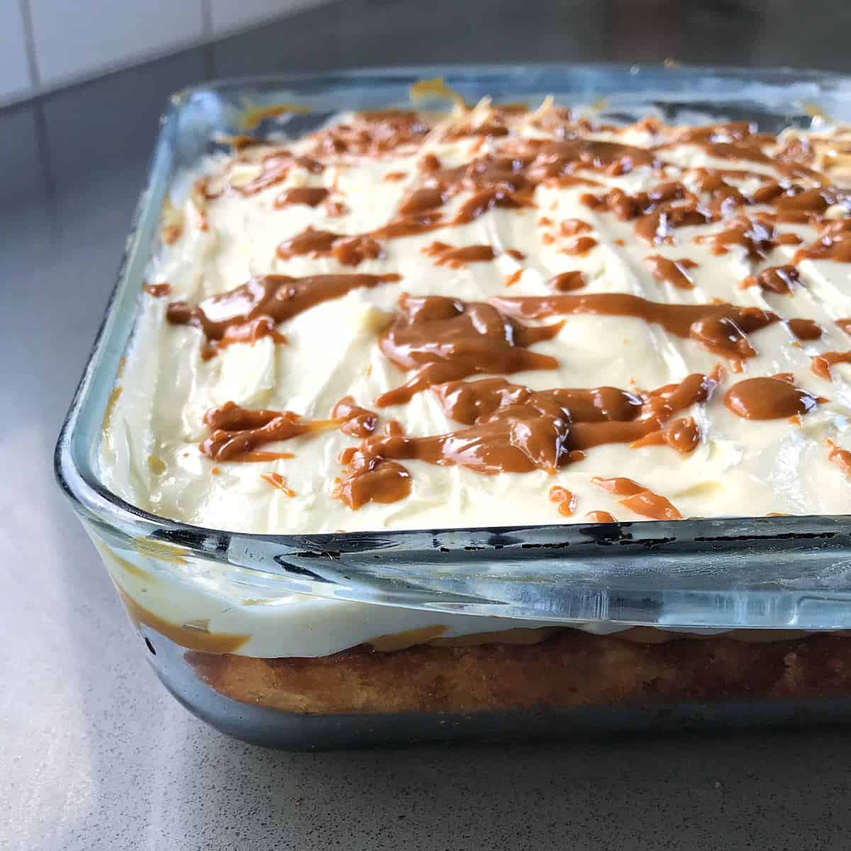A square glass baking dish containing a layered banana caramel cake topped with white frosting and drizzles of caramel sauce.