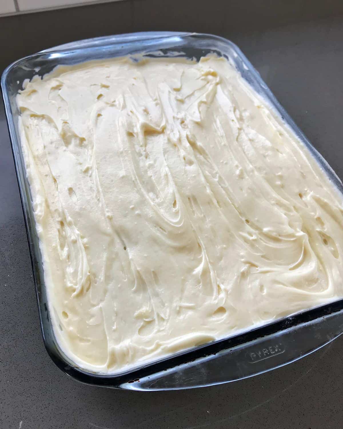 A rectangular glass baking dish filled with a banana caramel cake covered in thick, swirled white frosting.