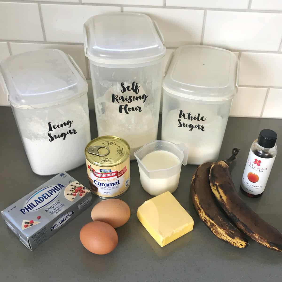 Baking ingredients for a banana caramel cake, including labeled storage containers, canned caramel, milk, eggs, butter, two overripe bananas, and vanilla extract.