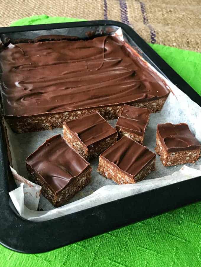 Dark Chocolate Peppermint Slice A tray of Dark Chocolate peppermint slice with a glossy chocolate topping, partially cut into square pieces, rests on parchment paper.