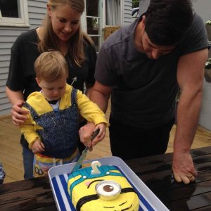 A family standing behind a minion cake that a boy is cutting into.