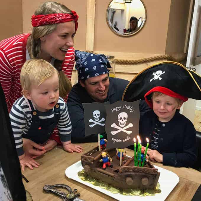 A family, dressed as pirates sitting around a boy with his pirate ship birthday cake.