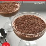 Two glass dessert dishes filled with rich chocolate mousse, topped with grated chocolate, are placed on a gray surface next to two spoons.