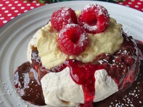 A delightful mini pavlova topped with cream, fresh raspberries, chocolate sauce, and powdered sugar, served on a plate—perfect for showcasing festive Mini Pavlovas at any celebration.