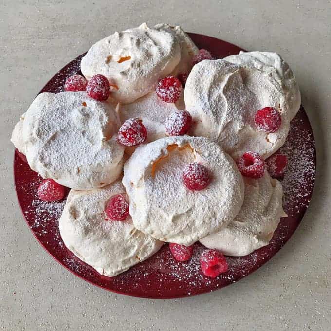 A red plate holds several mini Pavlovas dusted with powdered sugar and garnished with fresh raspberries.
