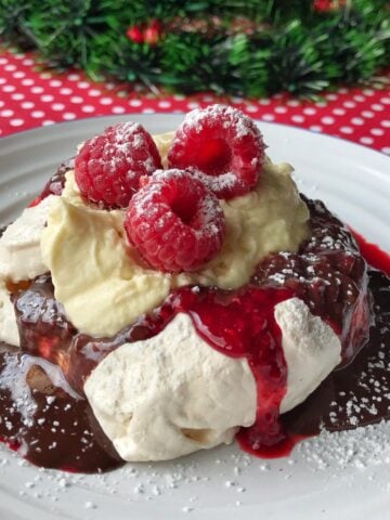 Mini Pavlovas topped with whipped cream, raspberry sauce, fresh raspberries, and powdered sugar are served on a white plate with festive decorations in the background.