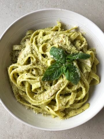 A white bowl filled with chicken pesto pasta-tender fettuccine coated in green pesto sauce, topped with grated cheese and a sprig of fresh basil.