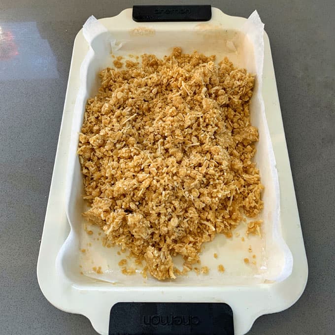 A baking dish lined with parchment paper holds an uneven layer of ginger crunch oat mixture, ready to be pressed or baked.