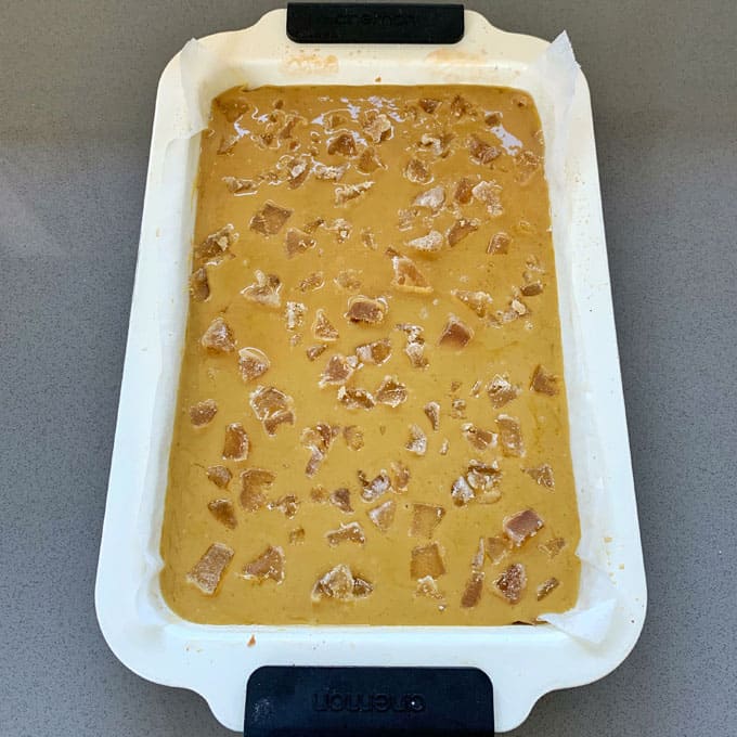 Rectangular white baking dish filled with unbaked ginger crunch batter, topped with small chunks, placed on a gray countertop.