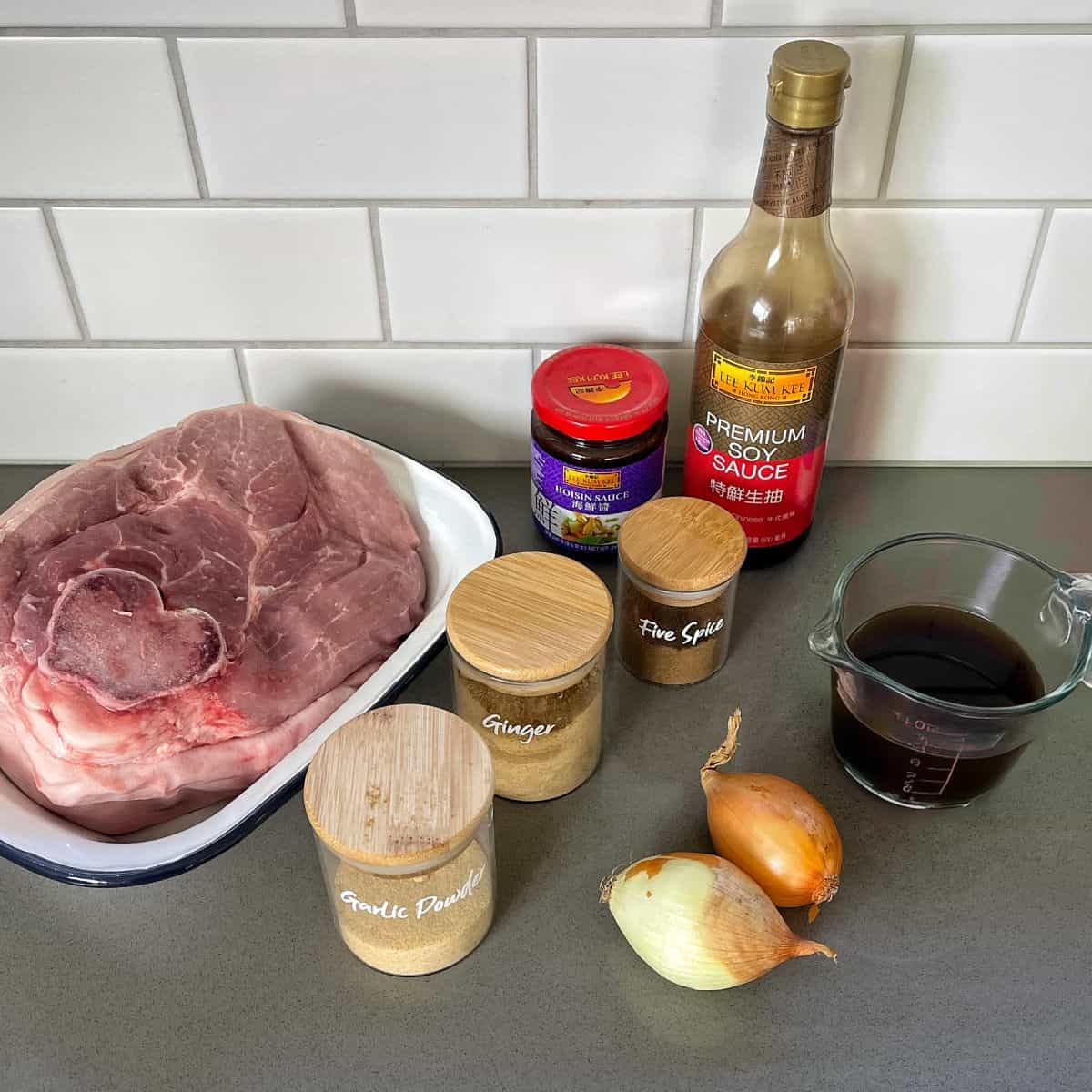 A piece of raw pork, soy sauce, jar of hoisin sauce, chili paste, garlic powder, ground ginger, five spice, and onions lay ready on the counter—perfect ingredients for making flavorful pulled pork.