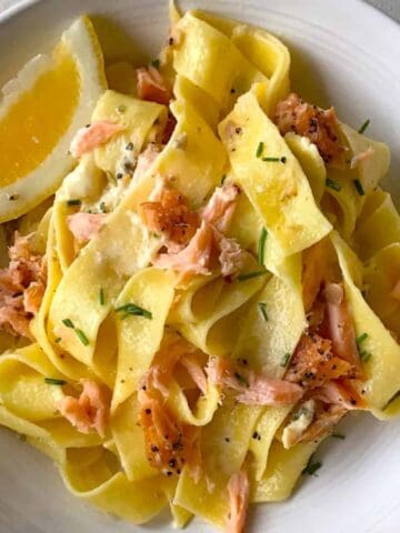 A bowl of wide ribbon pasta with flaked smoked salmon, chopped chives, black pepper, and a lemon wedge.