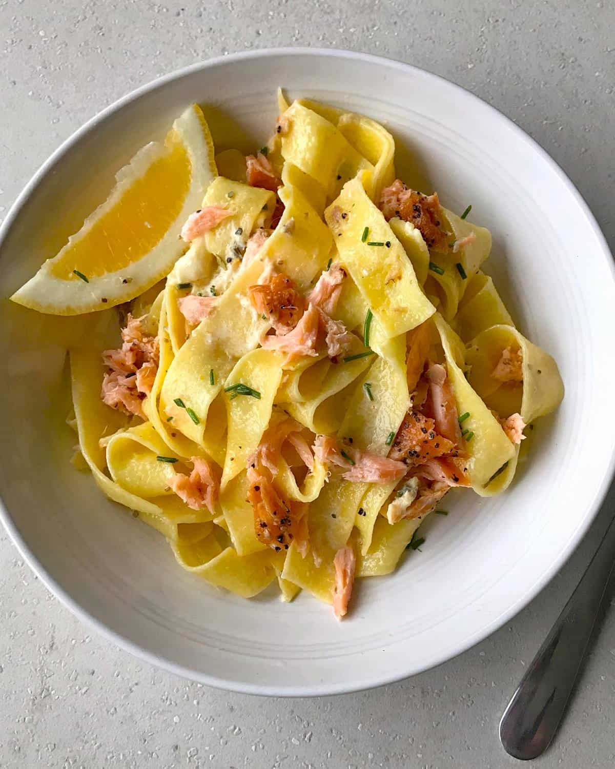VJ cooks smoked salmon pasta A white bowl of smoked salmon pasta featuring pappardelle with flaked salmon, chopped chives, black pepper, and a lemon wedge on the side. A metal spoon is placed next to the bowl.