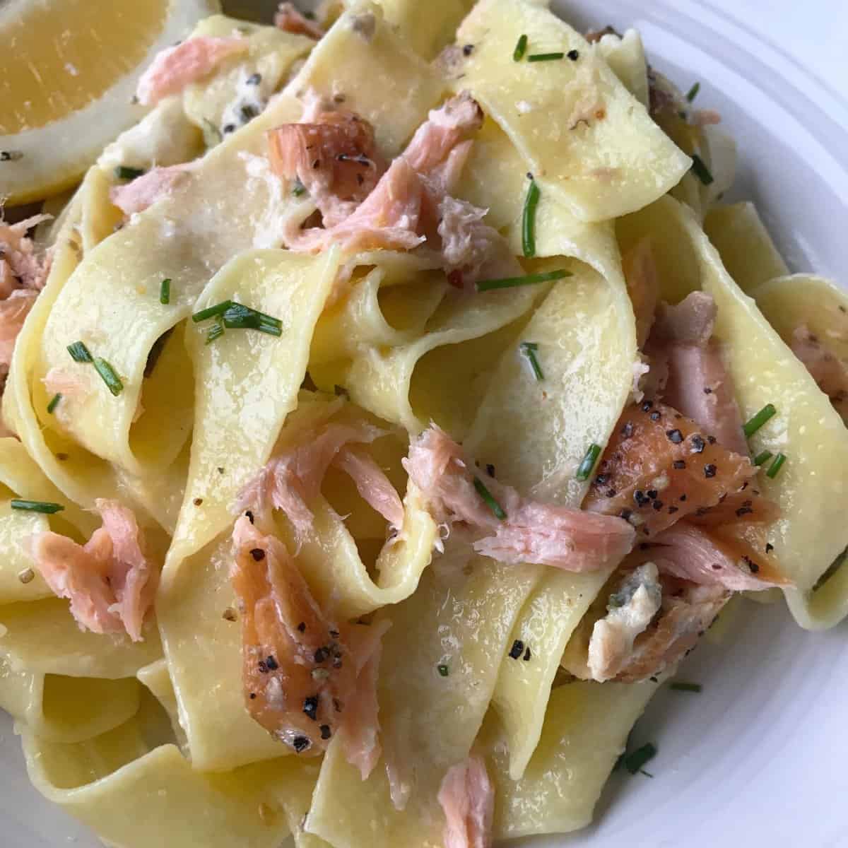 VJ cooks smoked salmon pasta Wide ribbon pasta topped with flaked smoked salmon, chopped chives, black pepper, and a lemon wedge on a white plate.