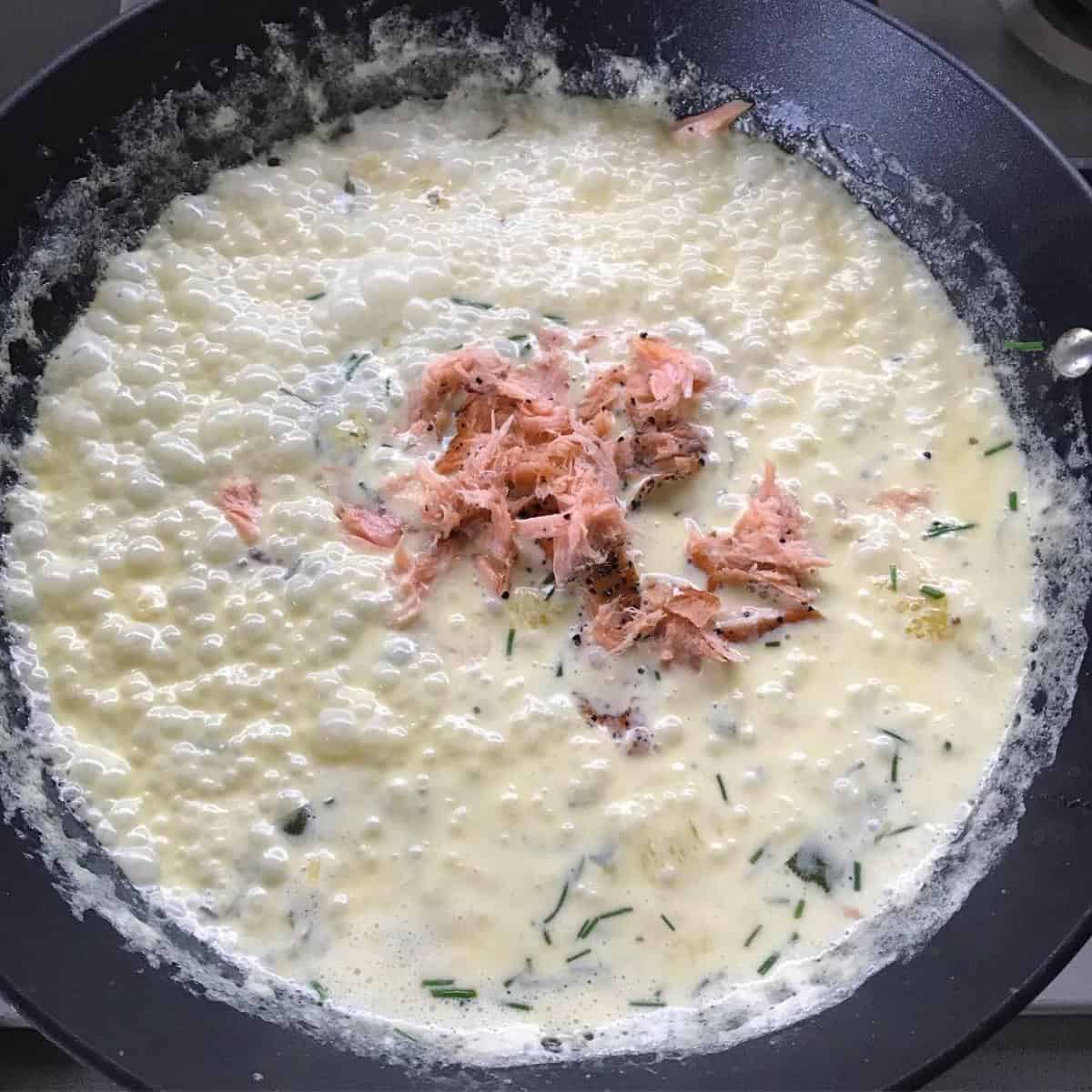 Smoked Salmon Pasta A creamy sauce with visible shredded salmon and chopped herbs simmers in a black skillet, creating the perfect base for smoked salmon pasta.