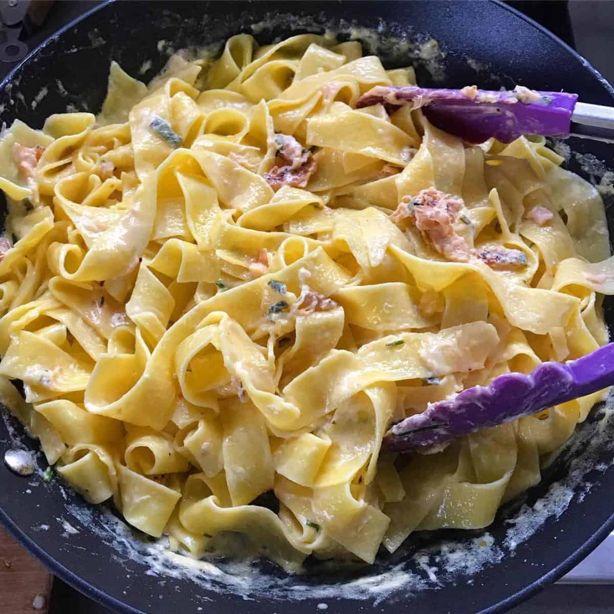 Smoked Salmon Pasta Wide ribbon pasta in a creamy yellow sauce with pieces of meat, being mixed in a pan with purple tongs—this dish is perfect for smoked salmon pasta lovers seeking comfort food with a gourmet twist.