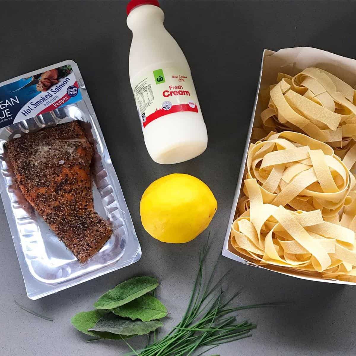 Smoked Salmon Pasta Smoked salmon, a bottle of fresh cream, a lemon, tagliatelle pasta, sage leaves, and chives are arranged on a gray surface—perfect ingredients for creating an elegant smoked salmon pasta dish.