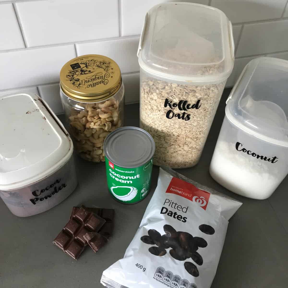 Raw Chocolate Slice Ingredients on a kitchen counter: cocoa powder, mixed nuts, rolled oats, coconut, coconut cream, pitted dates, and chocolate pieces—everything you need to create a delicious and healthy raw chocolate slice.