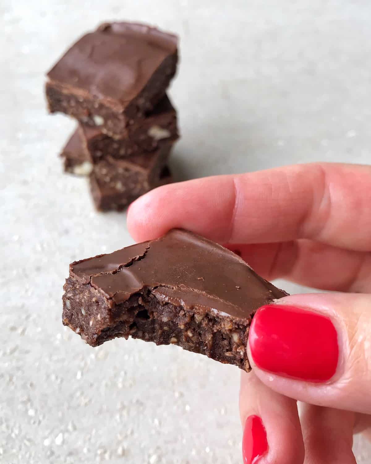 Raw Chocolate Slice A hand with red nail polish holds a square healthy raw chocolate slice with a bite taken out of it; more squares are blurred in the background.