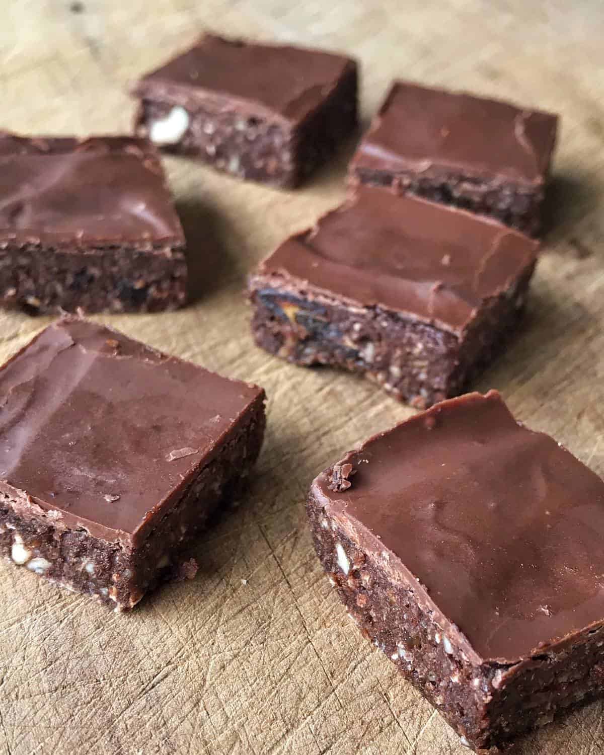 Six chocolate square bars with a glossy top layer are arranged on a wooden surface. These healthy raw chocolate slices have a dense, textured base with visible nut pieces.