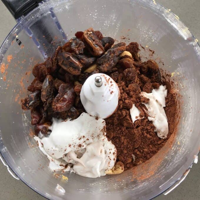 Raw Chocolate Slice A food processor bowl with dates, cocoa powder, cashews, raisins, and a white creamy ingredient before blending—perfect for making a healthy raw chocolate slice.