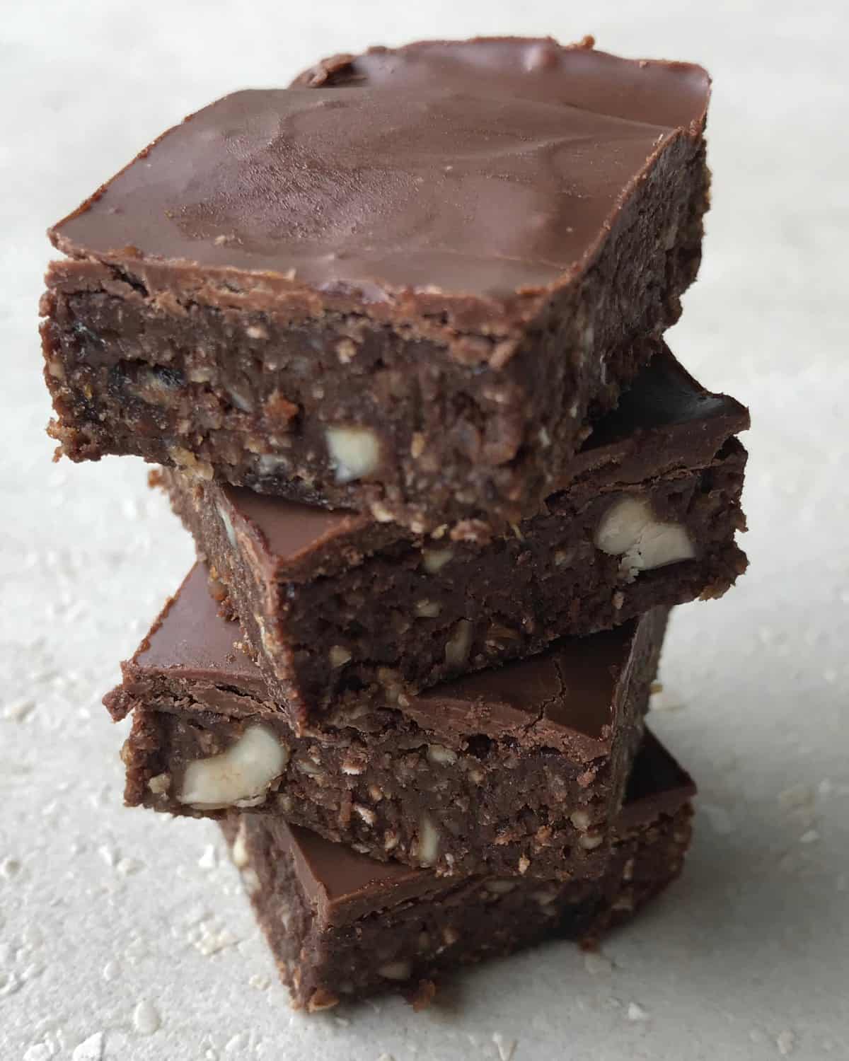 VJ cooks raw chocolate slice Three chocolate brownies with nuts are stacked on a light-colored surface, showing a dense, fudgy texture and a glossy chocolate topping reminiscent of a healthy raw chocolate slice.