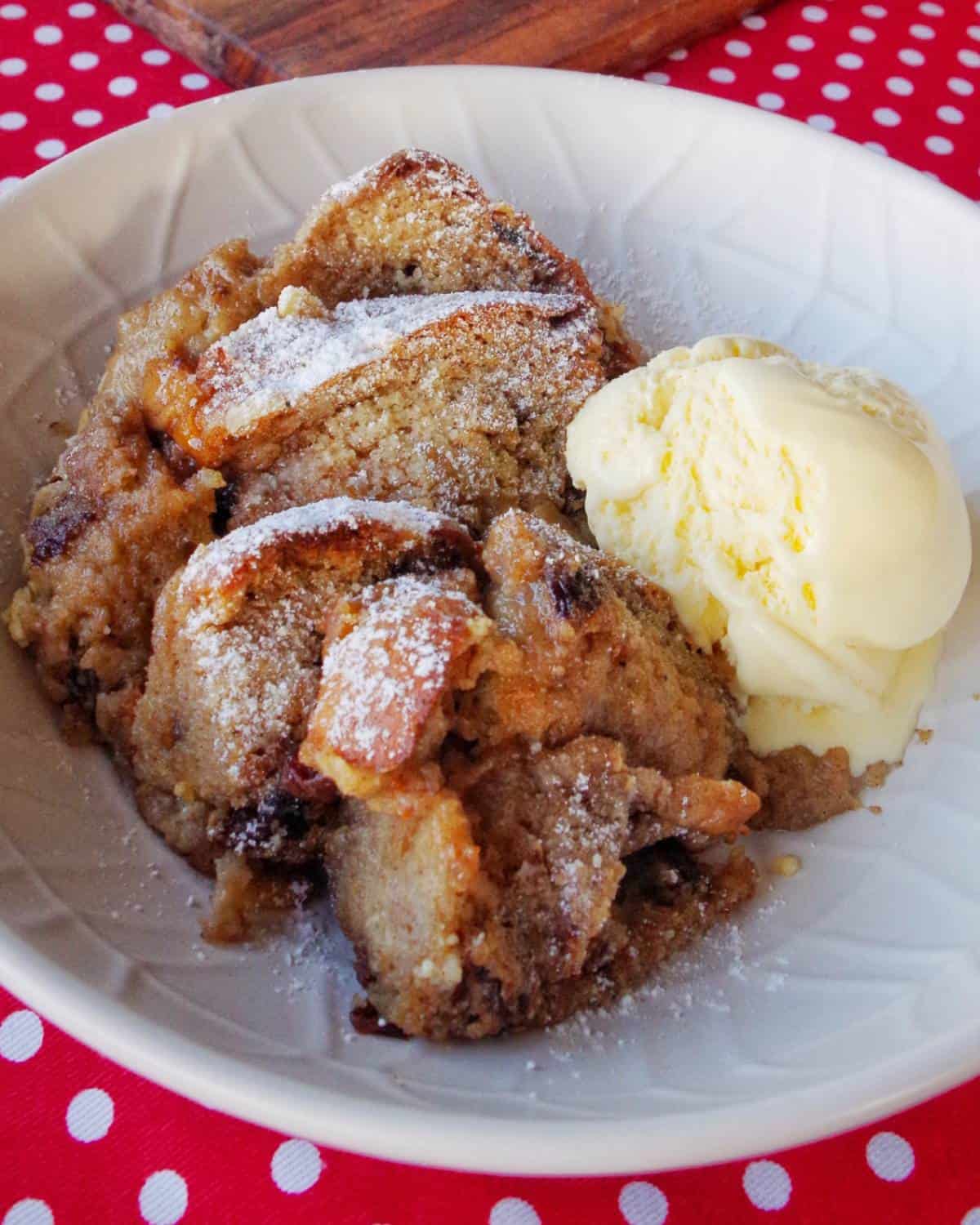 A bowl of hot cross bun bread and butter pudding topped with powdered sugar, served alongside a scoop of vanilla ice cream on a red polka dot tablecloth.