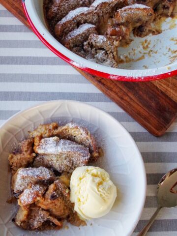 A bowl of Hot Cross Bun Bread and Butter Pudding with a scoop of vanilla ice cream sits next to a partially eaten serving in a red dish on a striped cloth.