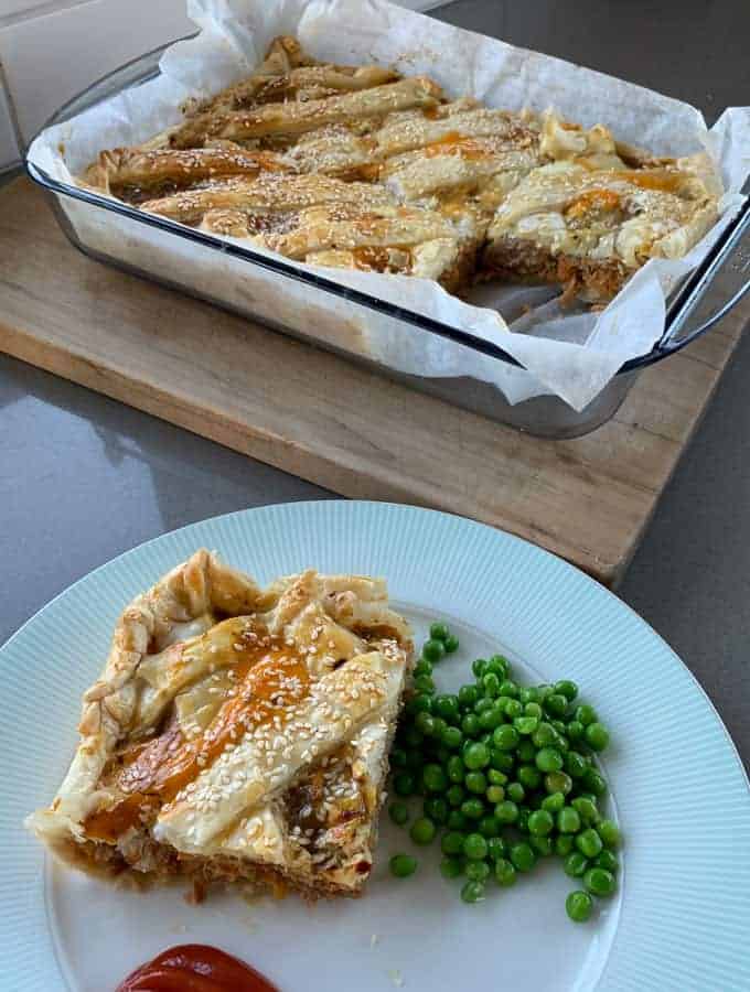 Sausage Picnic Pie A slice of savory sausage pie with golden pastry and sesame seeds is served on a plate with green peas and ketchup; the rest of the pie sits in a baking dish behind.