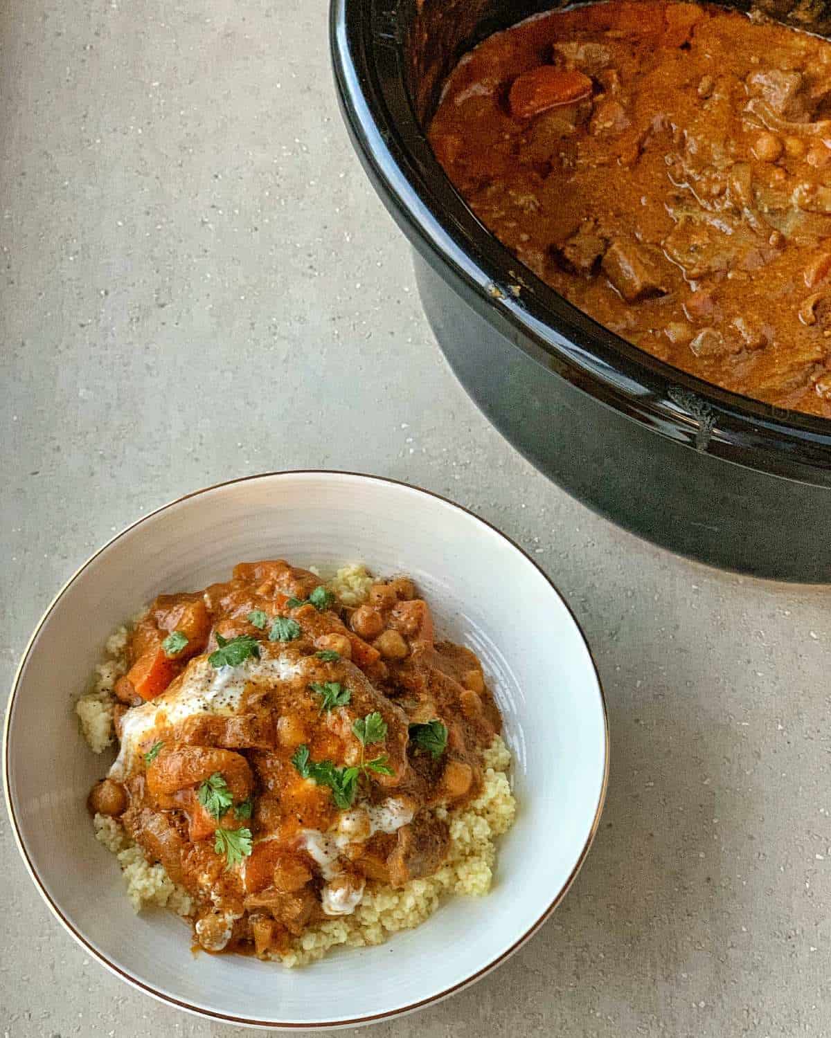 Slow Cooker Moroccan Lamb with Apricots A bowl of cooked grains topped with a saucy slow cooker Moroccan lamb stew filled with vegetables and herbs, next to a slow cooker brimming with more of the flavorful stew.
