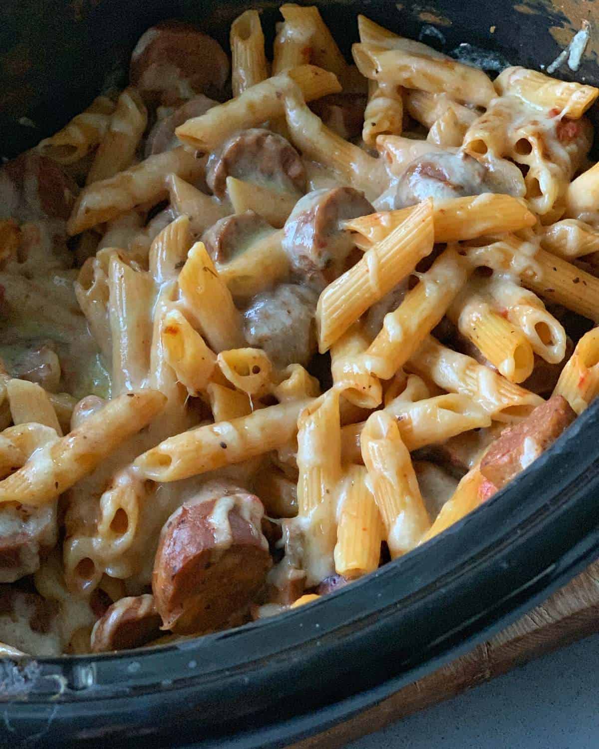 Slow cooker sausage pasta featuring penne, sliced sausage, and melted cheese in a black slow cooker.