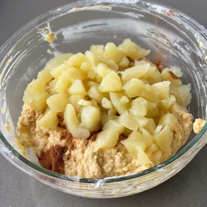 A glass bowl containing batter for oaty muffins with a heap of chopped apples and a sprinkle of cinnamon on top, set on a gray countertop.