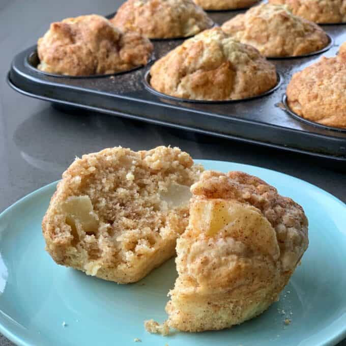 Easy Oaty Muffins A sliced oaty muffin with visible apple chunks sits on a blue plate, with a tray of whole muffins in the background.