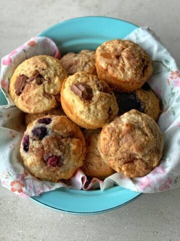 A blue bowl lined with a floral cloth holds assorted muffins, including oaty muffins with chocolate pieces and others bursting with berries.