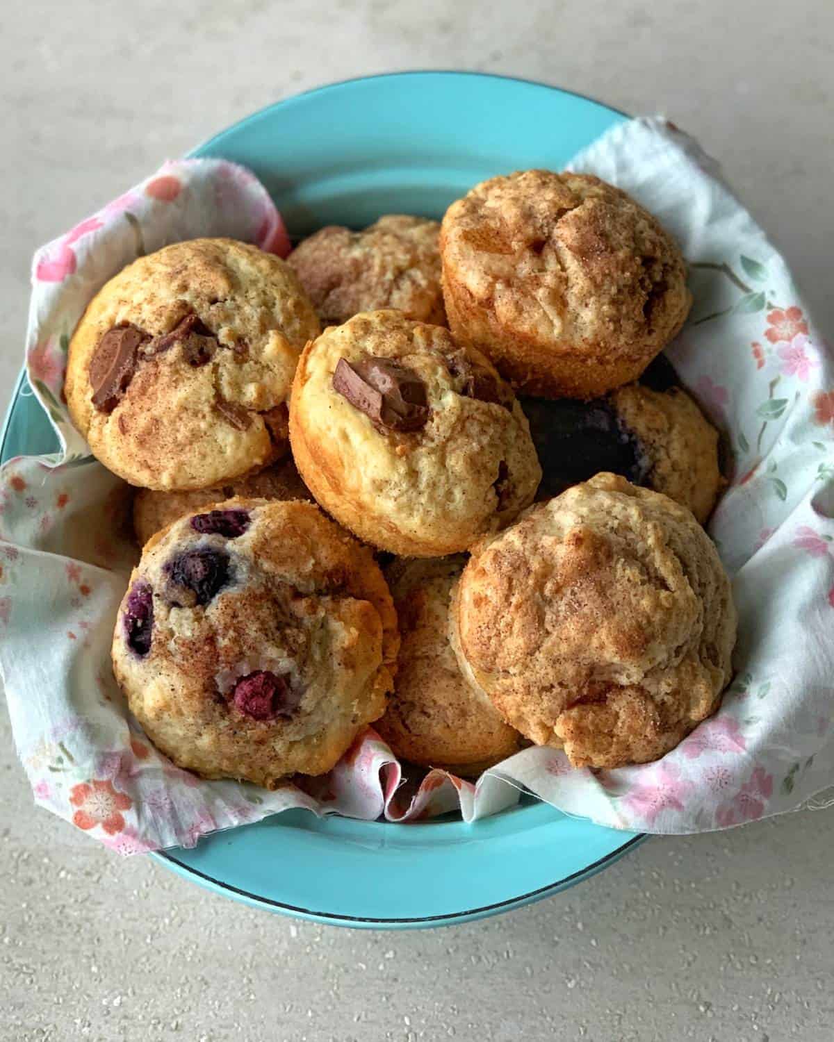 A blue bowl lined with a floral cloth holds assorted muffins, including oaty muffins with chocolate pieces and others bursting with berries.