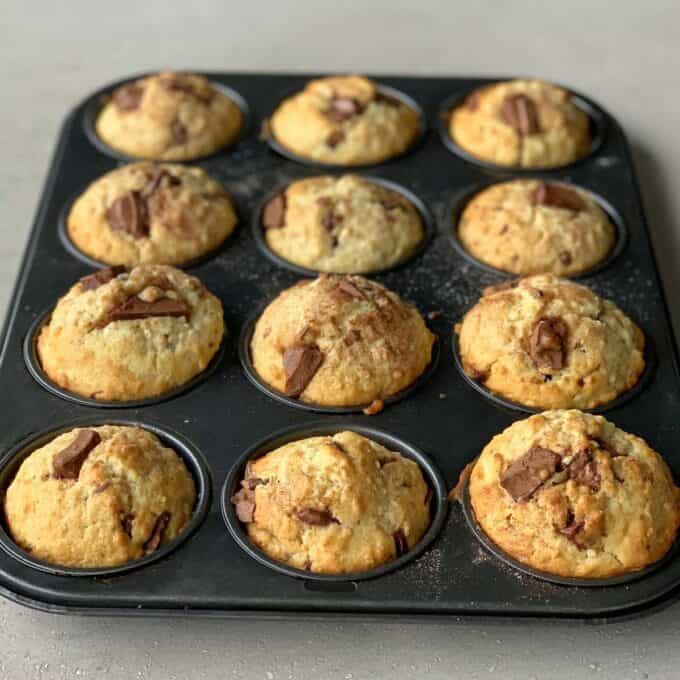 A muffin tray with twelve freshly baked oaty muffins, each topped with pieces of chocolate, on a light-colored surface.