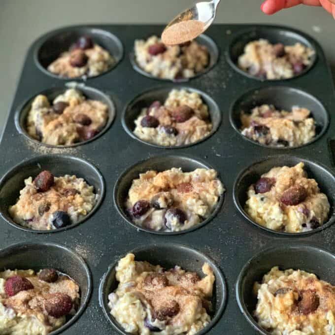 Easy Oaty Muffins A hand sprinkles cinnamon sugar onto unbaked oaty muffin batter, studded with blueberries, in a twelve-cup muffin pan.
