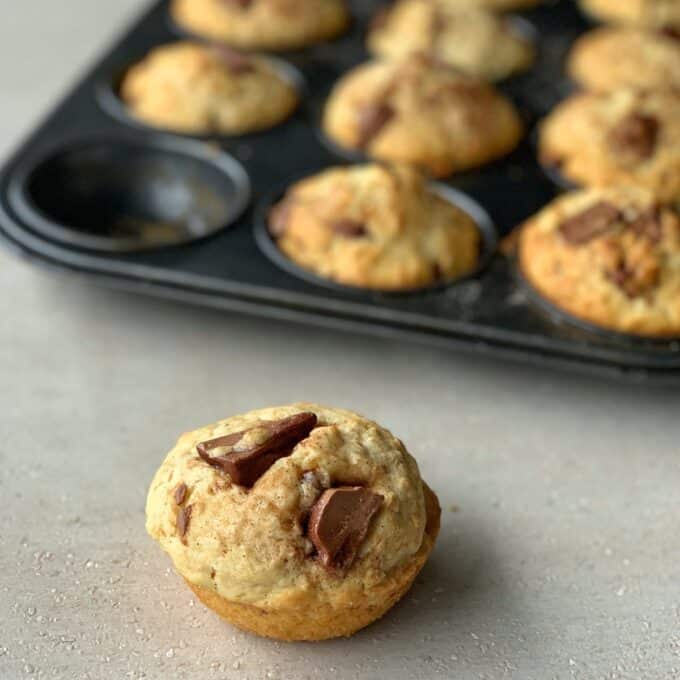 A close-up of a chocolate chip muffin on a counter, with a muffin tin holding more oaty muffins in the background.
