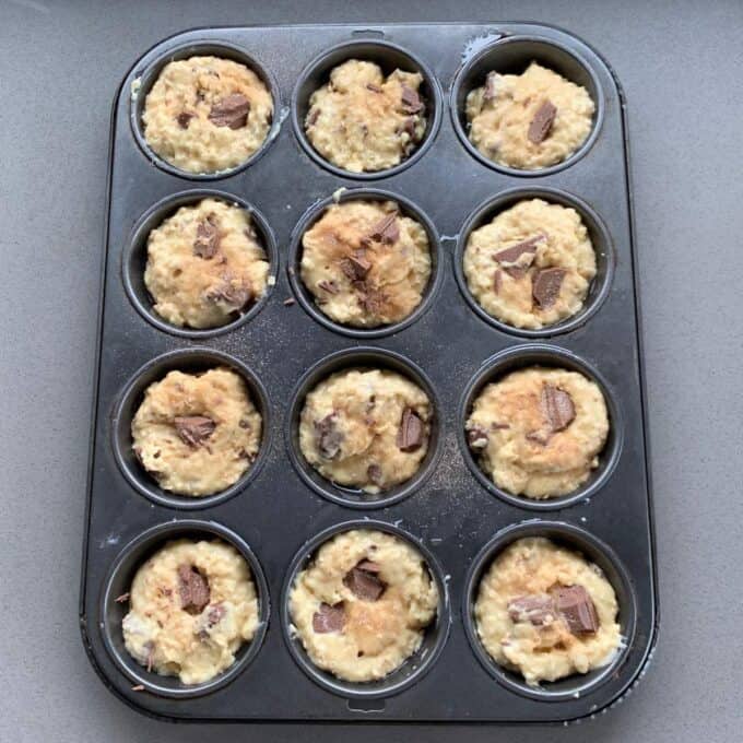 A muffin tin filled with unbaked oaty muffin batter containing visible chocolate chunks, placed on a gray countertop.