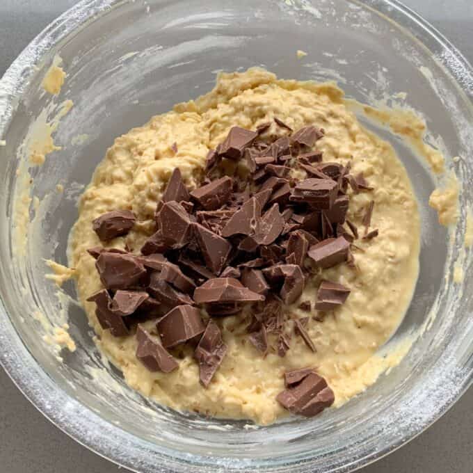 A glass bowl containing a thick, pale batter for oaty muffins, with chunks of milk chocolate scattered on top, ready to be mixed in.