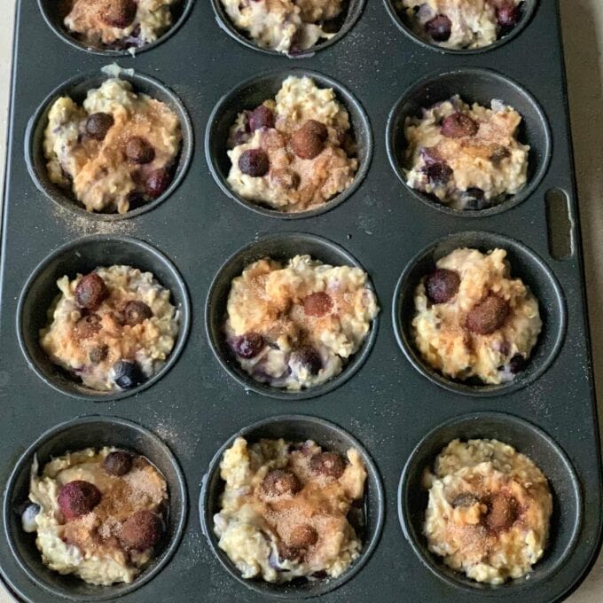 Easy Oaty Muffins A muffin tin filled with unbaked oaty muffins batter containing blueberries and sprinkled with cinnamon and sugar, ready to be placed in the oven.