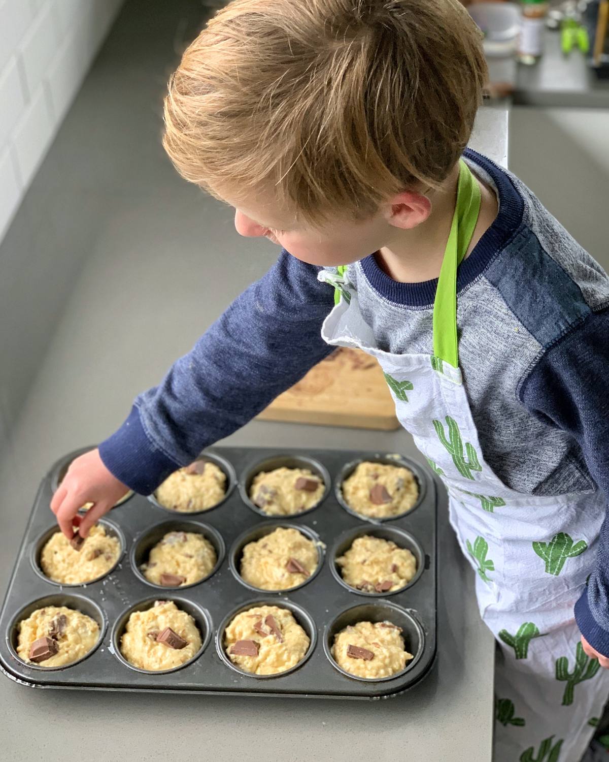 A young child in a cactus-patterned apron adds toppings to oaty muffins batter in a muffin tin on a kitchen counter.