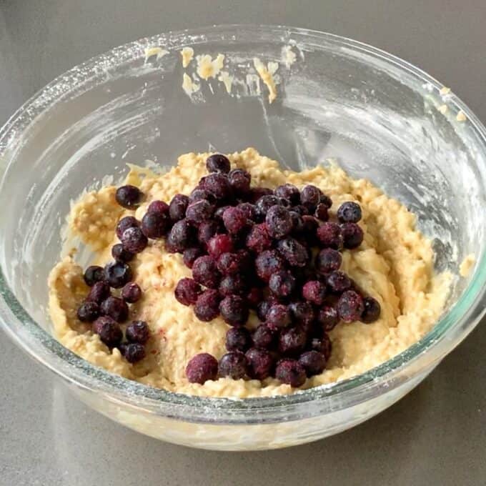 Easy Oaty Muffins A glass bowl filled with oaty muffin batter and a pile of blueberries on top, sitting on a gray countertop.