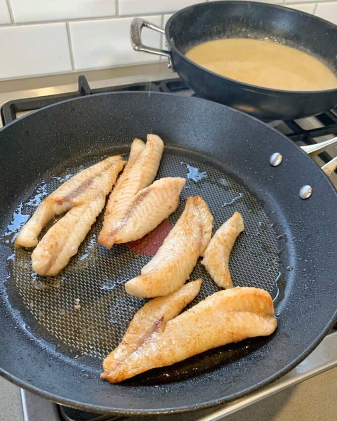 Several fillets of fish are being pan-fried in a black skillet, while a second pan in the background simmers a light-colored Thai green curry sauce.