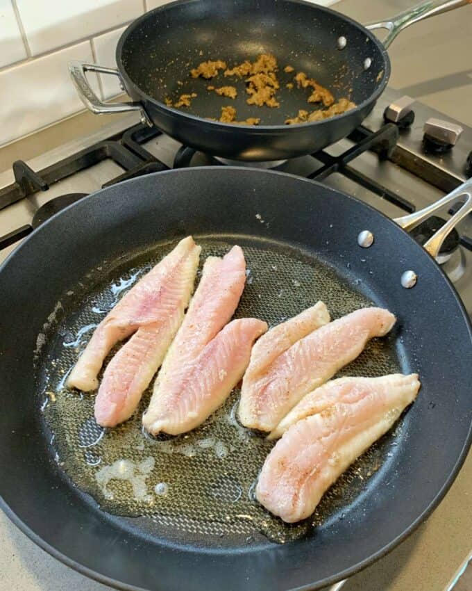 Five raw fish fillets are cooking in oil in a frying pan on a stove, perfect for making pan fried fish with Thai green curry sauce. Another pan with cooked residue sits on a different burner in the background.