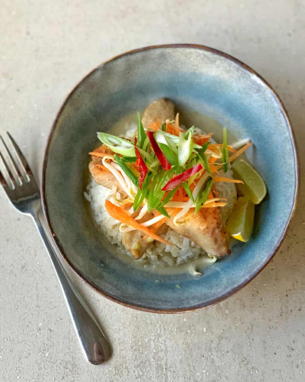 VJ cooks - Pan fried fish with Thai green curry sauce A bowl of rice topped with pan fried fish with Thai green curry sauce, julienned carrots, green onions, red chili, and lime wedges, with a fork placed beside the bowl.