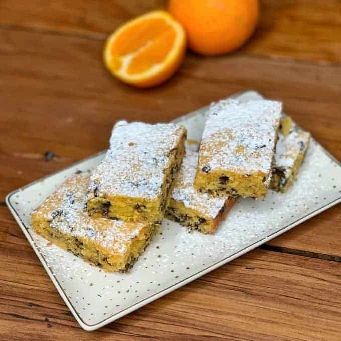 Rectangular pieces of orange blondie with dark chocolate chips, dusted with powdered sugar, rest on a white plate, with a halved orange and a whole orange in the background.